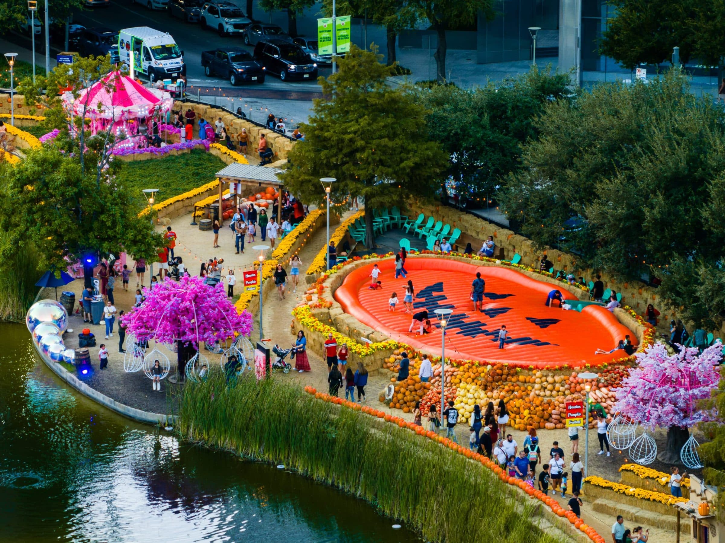 Aerial view of the Houston Pumpkin Festival at Discovery Green, with the giant inflatable jack-o'-lantern, pink blossom trees, carousel and downtown Houston streets behind