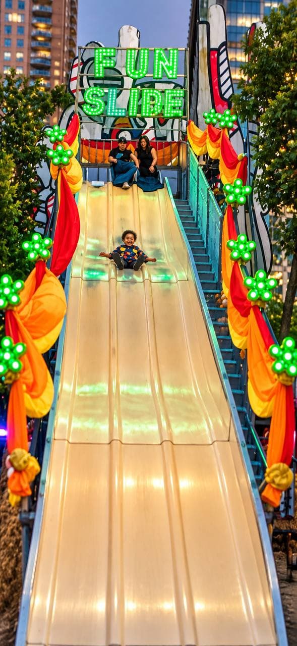 A grinning child racing down the 30-foot fun slide under its glowing green sign, with more riders waiting at the top